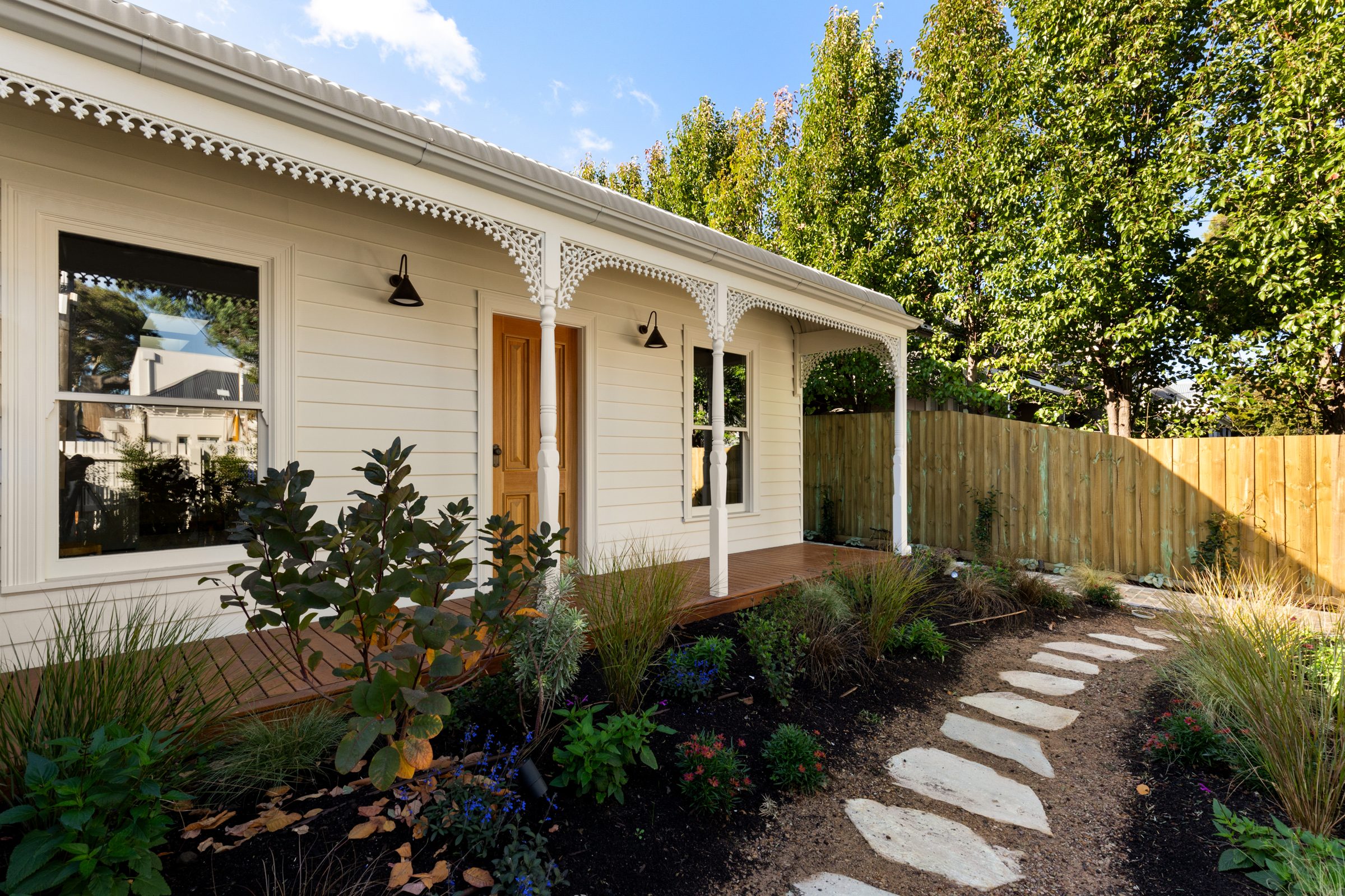The front verandah with timber decking, fretwork and turned posts, a planted garden and stepping-stone path beside a timber fence